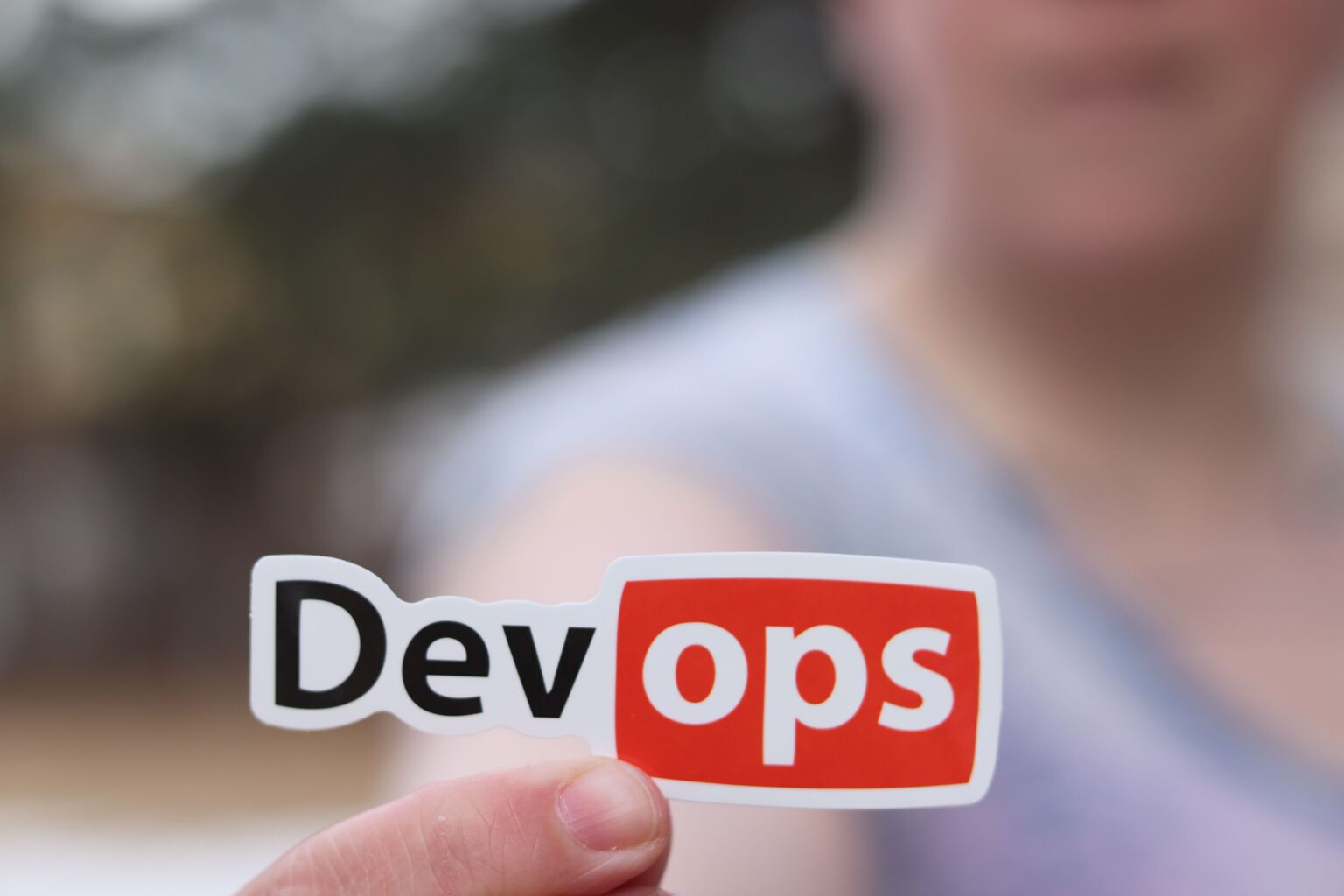 A hand holds a sticker with the word DevOps on it.